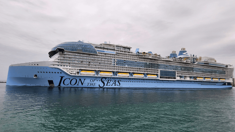 A cruise ship moored at a dock, reflecting in the water under a bright blue sky.