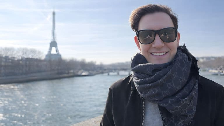 Zach Randles-Friedman smiling outdoors near the Seine River with the Eiffel Tower in the background, wearing sunglasses and scarf.