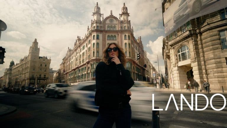 Person in sunglasses stands confidently on a busy city street, with ornate historic building in the background. Motion blur from passing cars adds dynamic energy.