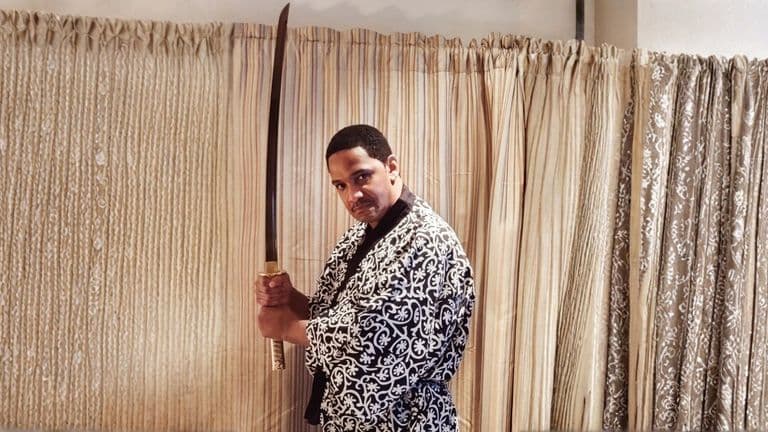 A person in a patterned kimono holds a katana upright with both hands, in front of a curtain backdrop. The expression is focused and intense.