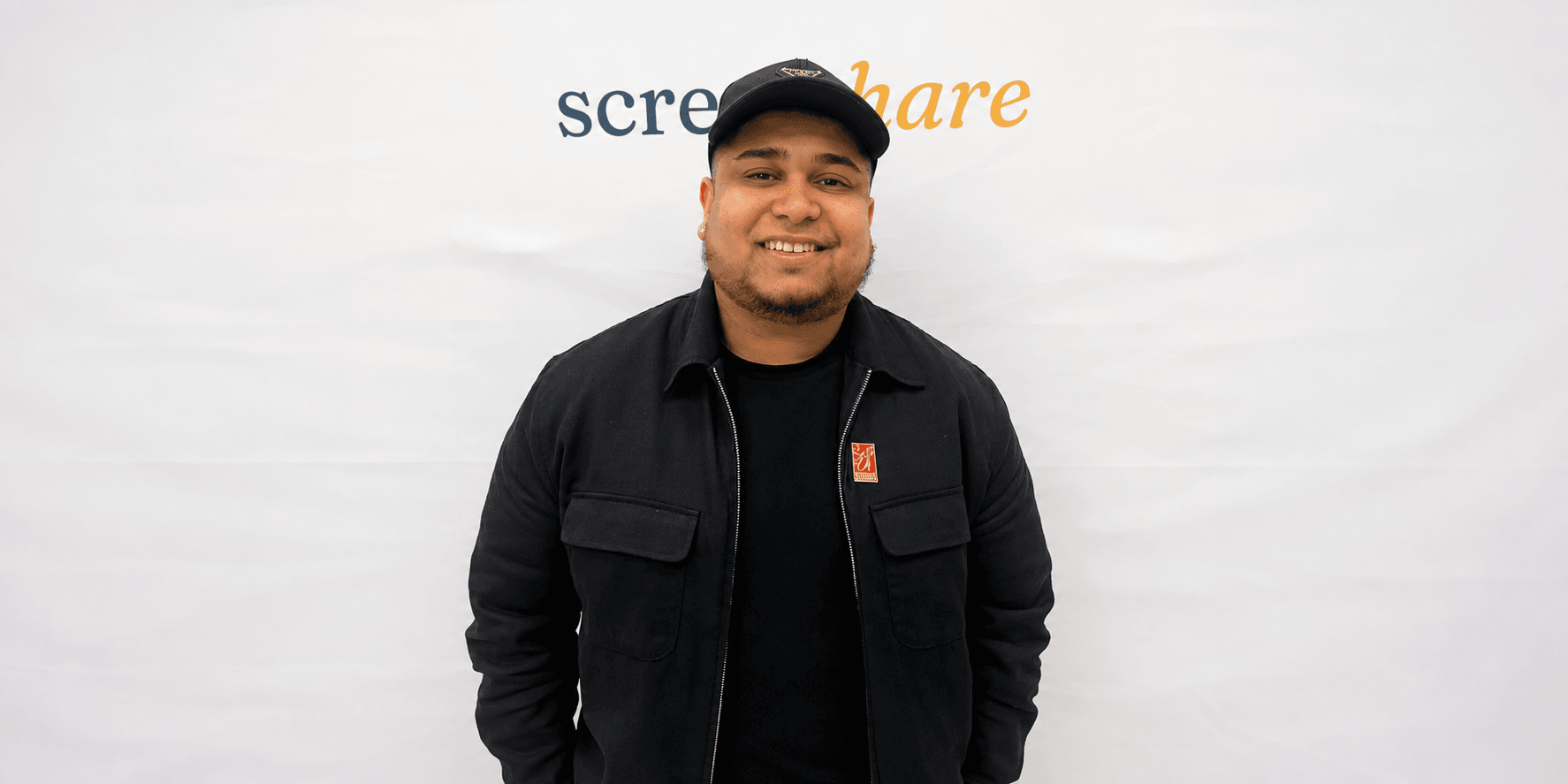 Smiling person in black jacket and cap stands in front of a white backdrop
