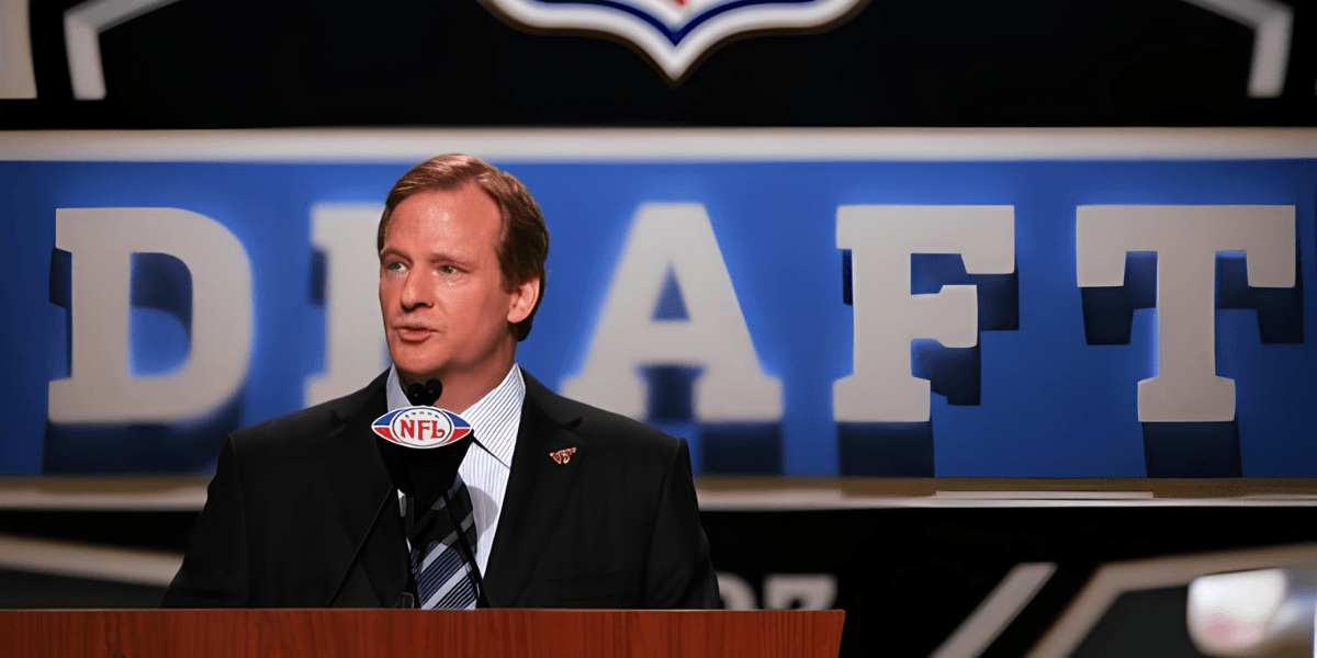 Man in a suit speaking at NFL Draft event with large "DRAFT" sign behind him