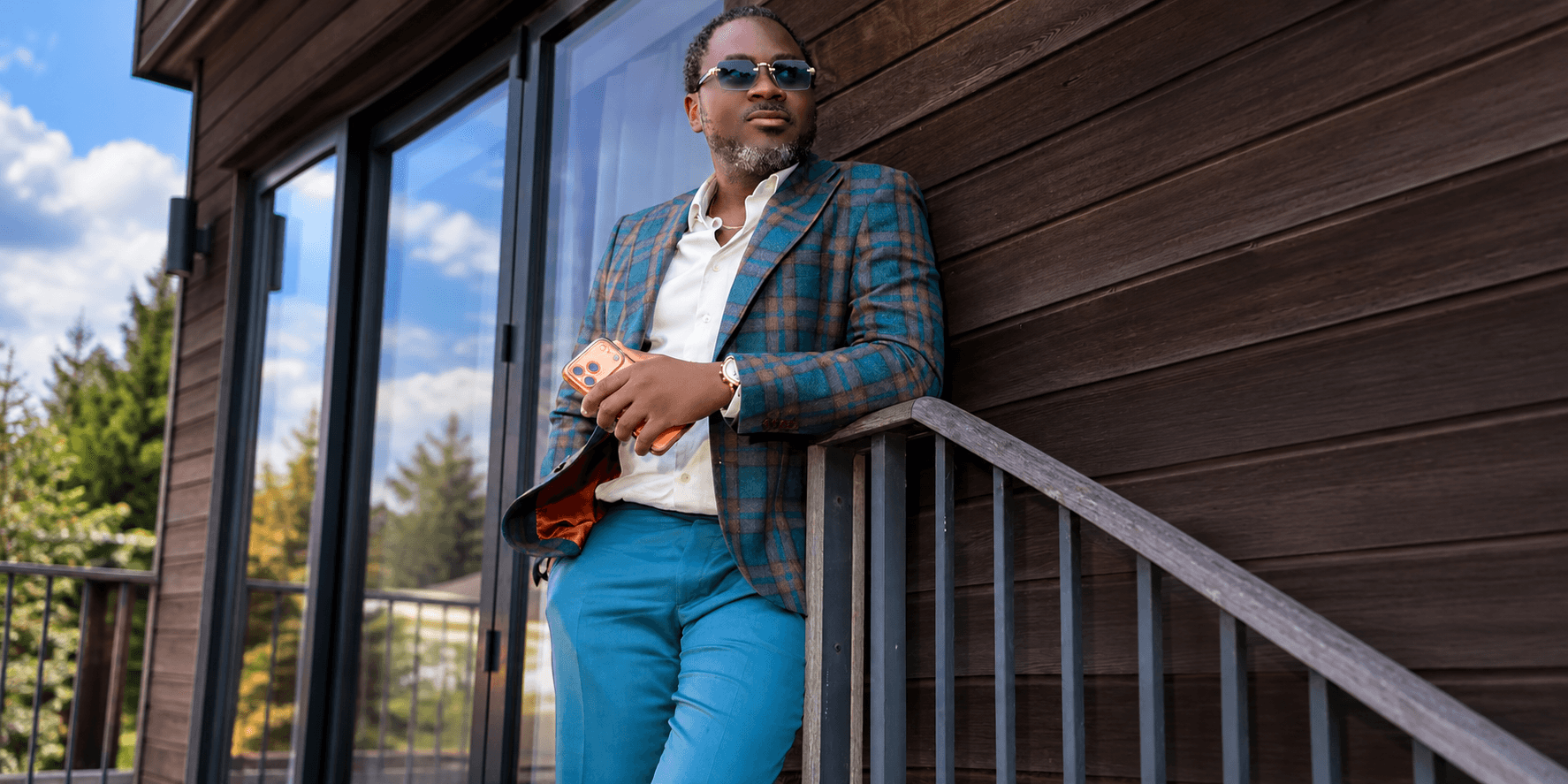 A man in a patterned blazer and sunglasses leans against a wooden railing