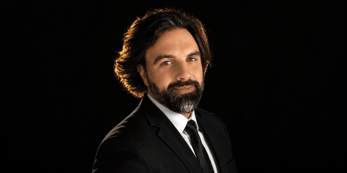 Man in a black suit with a beard, smiling against a dark background