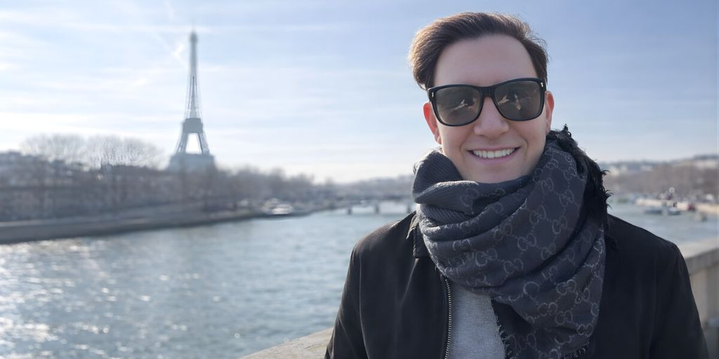Zach Randles-Friedman smiling outdoors near the Seine River with the Eiffel Tower in the background, wearing sunglasses and scarf.