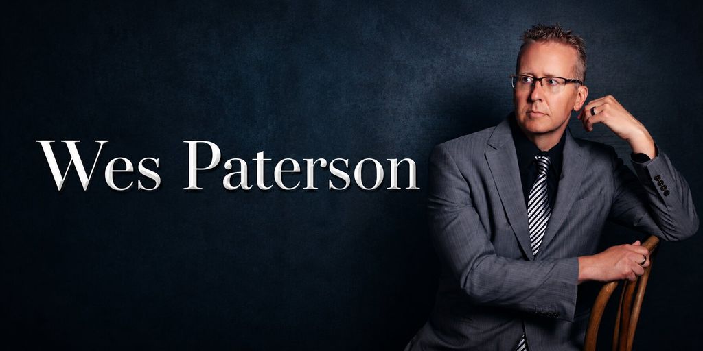 Portrait of Wesley Paterson in a gray suit and glasses against a dark background.