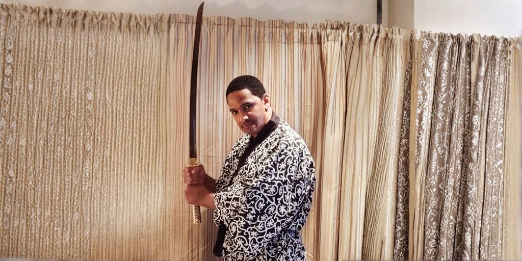 A person in a patterned kimono holds a katana upright with both hands, in front of a curtain backdrop. The expression is focused and intense.