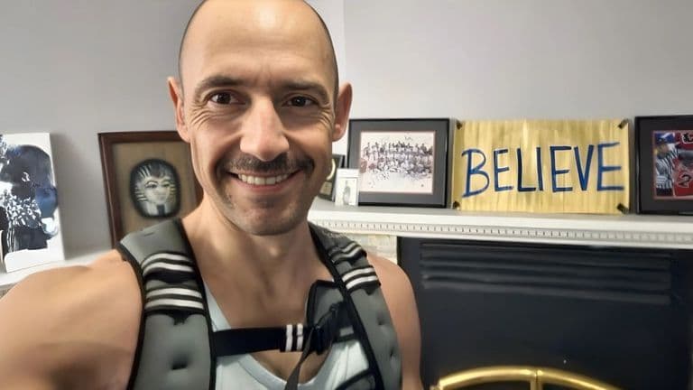 Motivational photo of a person wearing a weighted vest, standing in front of a fireplace with framed artwork and a "BELIEVE" sign on the wall. The person smiles confidently, emphasizing strength and perseverance. The background features a mix of inspiring art pieces and a personal sports memorabilia display.