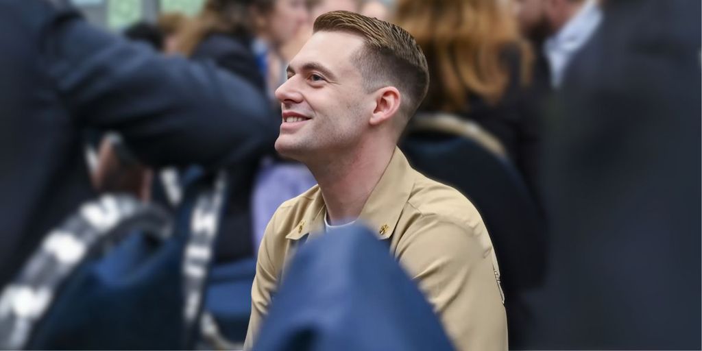 Smiling man in a beige uniform seated in a conference or event setting, surrounded by people, with a focused and engaging expression.