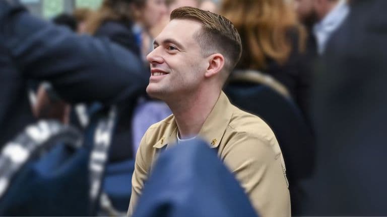 Smiling man in a beige uniform seated in a conference or event setting, surrounded by people, with a focused and engaging expression.
