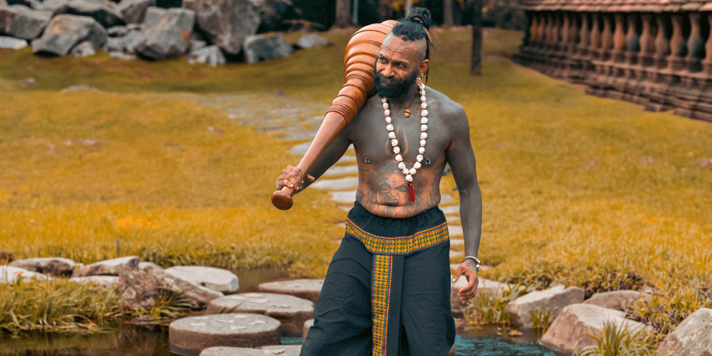A muscular man with tattoos carries a large wooden club in a serene landscape