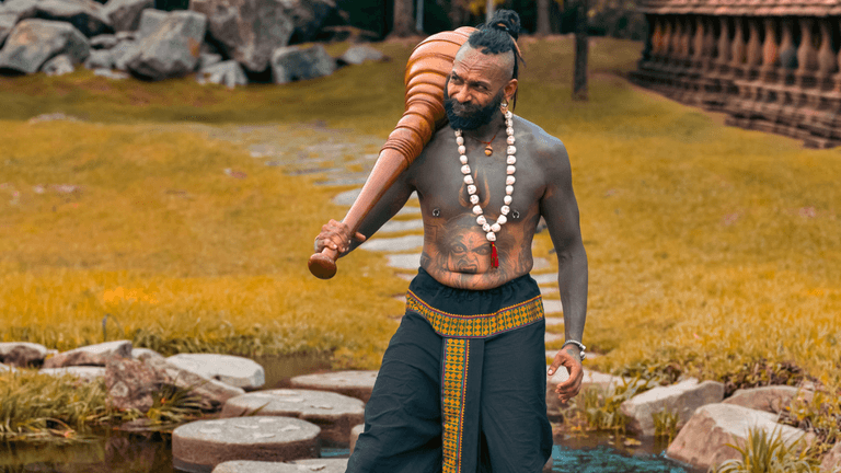 A muscular man with tattoos carries a large wooden club in a serene landscape
