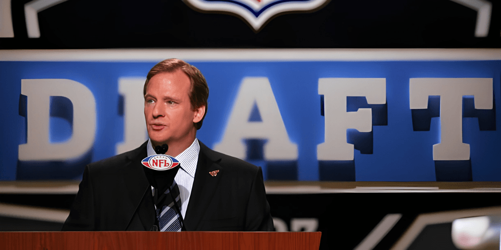Man in a suit speaking at NFL Draft event with large "DRAFT" sign behind him