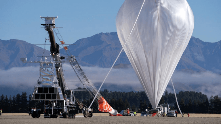 A large balloon being prepared for launch alongside a scientific equipment platform in a mountainous area