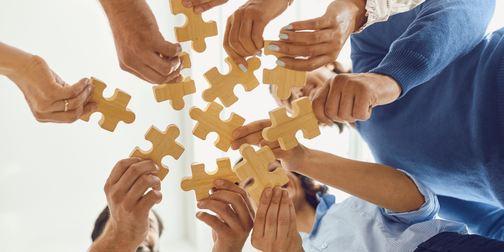 Multiple hands reaching for wooden puzzle pieces in a collaborative, team-building setting