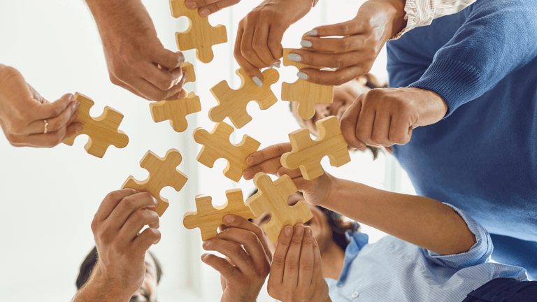 Multiple hands reaching for wooden puzzle pieces in a collaborative, team-building setting