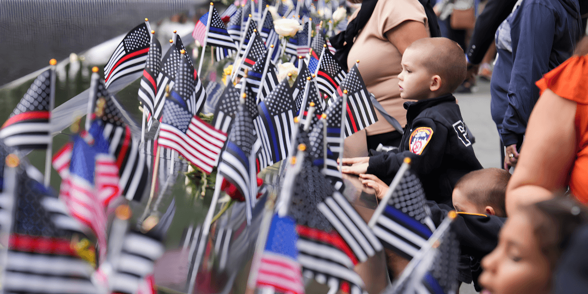 Children and adults observe flags and flowers in a memorial setting.