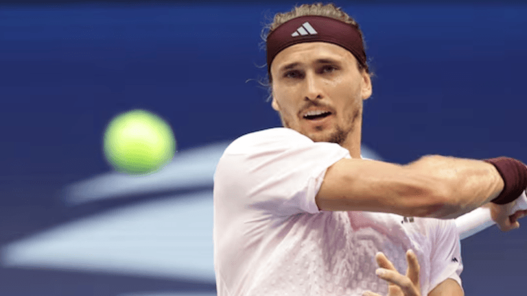 A male tennis player with long hair swings his racket at a ball.