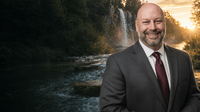 A smiling man in a suit stands by a scenic waterfall and river backdrop