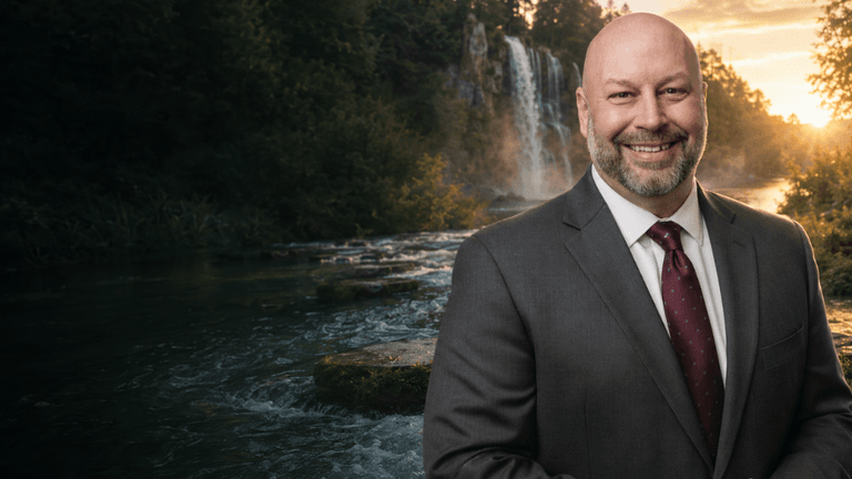 A smiling man in a suit stands by a scenic waterfall and river backdrop