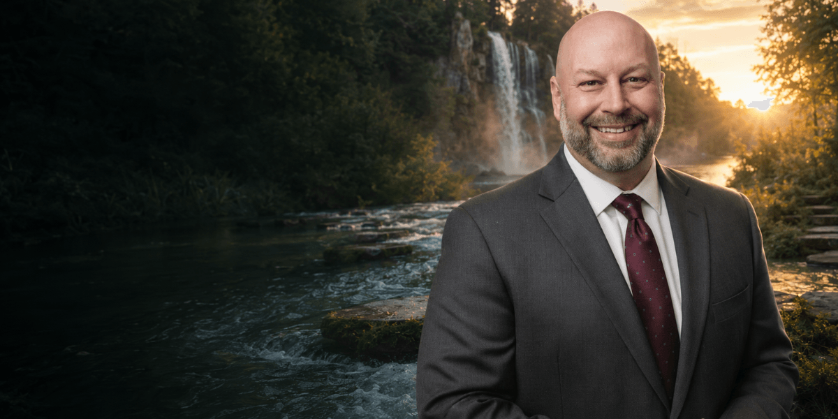 A smiling man in a suit stands by a scenic waterfall and river backdrop