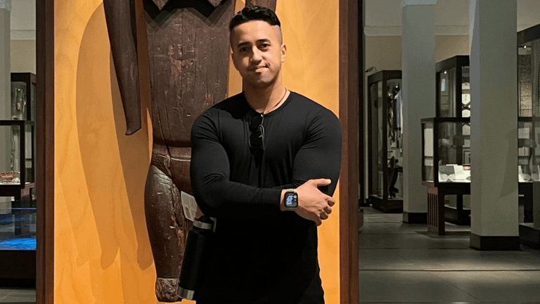 Man in a museum posing beside a large wooden artifact wearing a black shirt