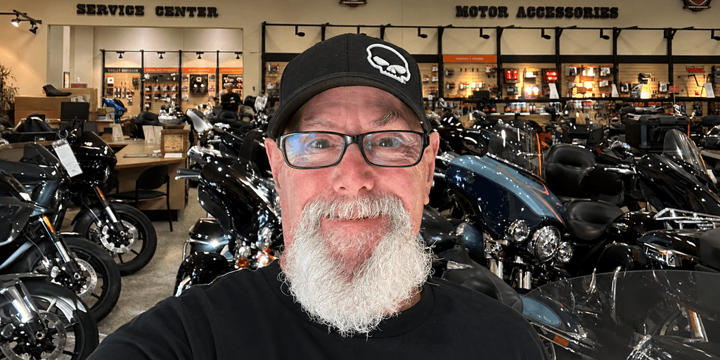 Man with a beard and glasses smiles in a motorcycle showroom full of bikes