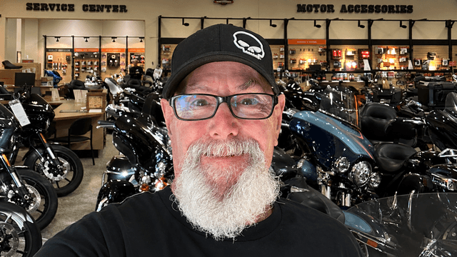 Man with a beard and glasses smiles in a motorcycle showroom full of bikes