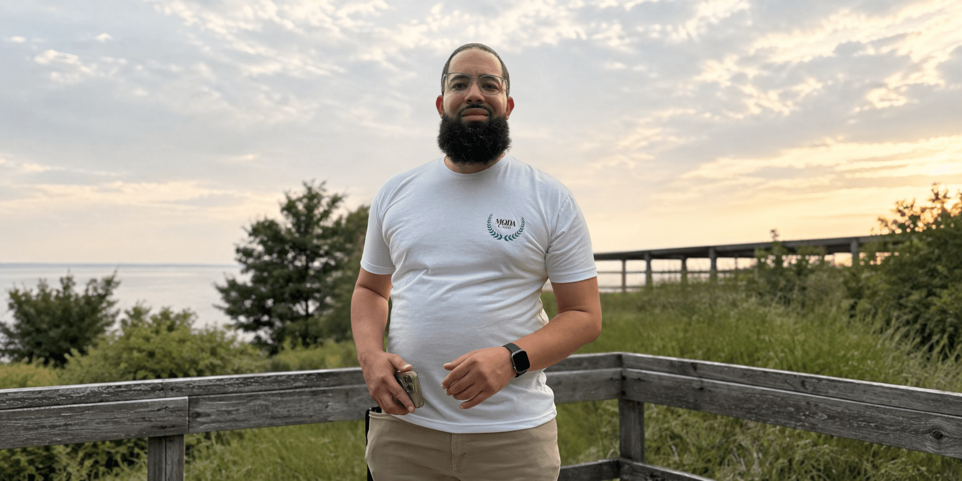 Man standing outdoors by a waterfront during sunset, wearing a white t-shirt and beige pants