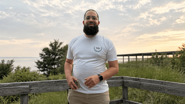 Man standing outdoors by a waterfront during sunset, wearing a white t-shirt and beige pants