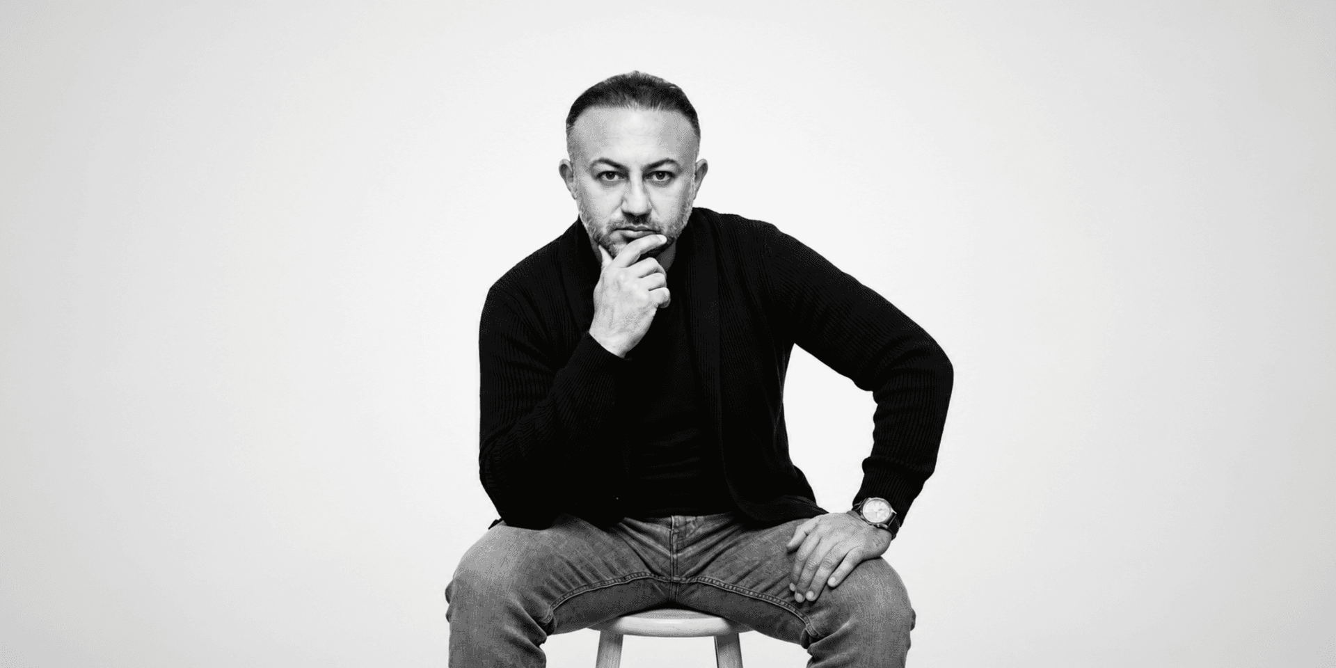 A man with a thoughtful expression sits on a stool, wearing casual clothing.