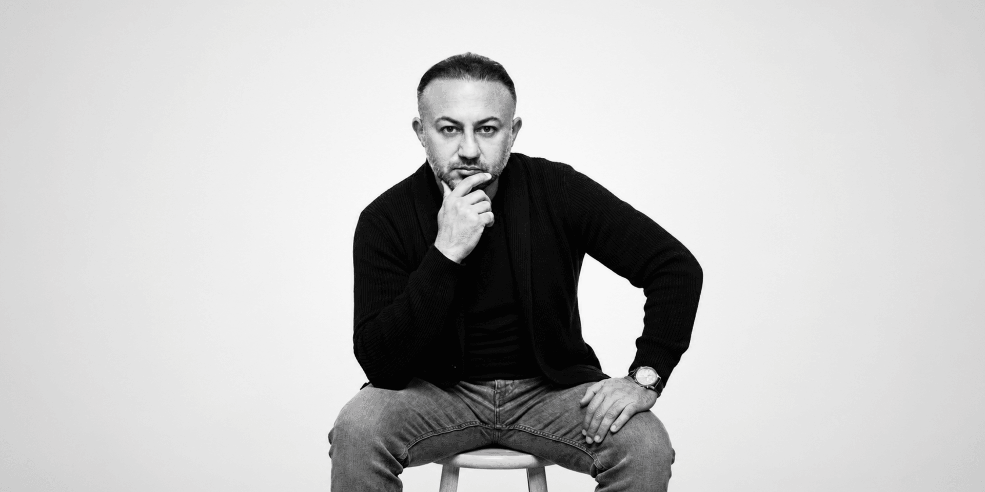 A man with a thoughtful expression sits on a stool, wearing casual clothing.