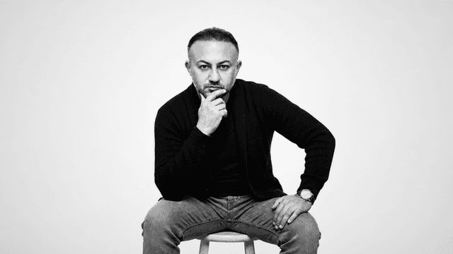 A man with a thoughtful expression sits on a stool, wearing casual clothing.