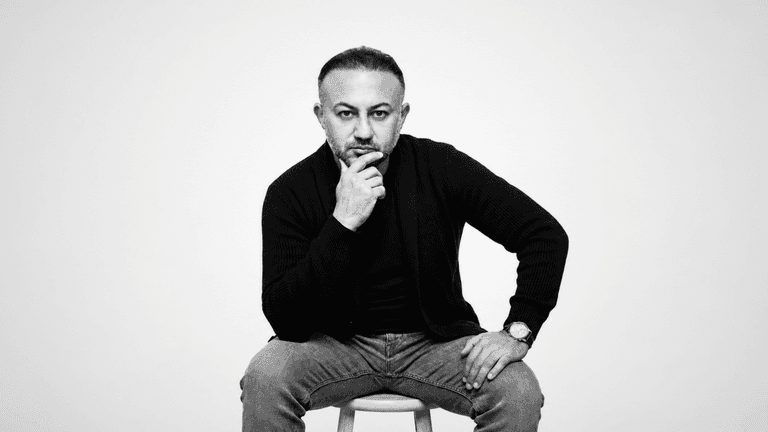 A man with a thoughtful expression sits on a stool, wearing casual clothing.
