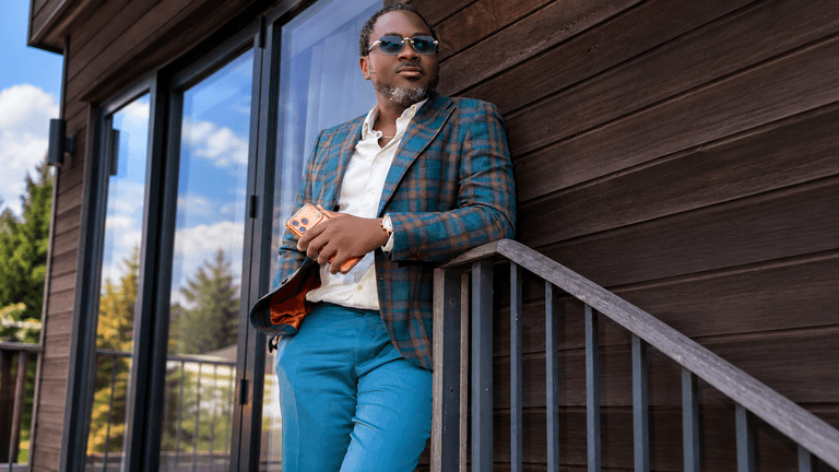 A man in a patterned blazer and sunglasses leans against a wooden railing