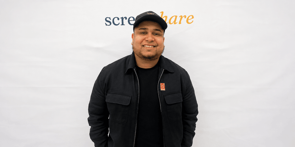 Smiling person in black jacket and cap stands in front of a white backdrop