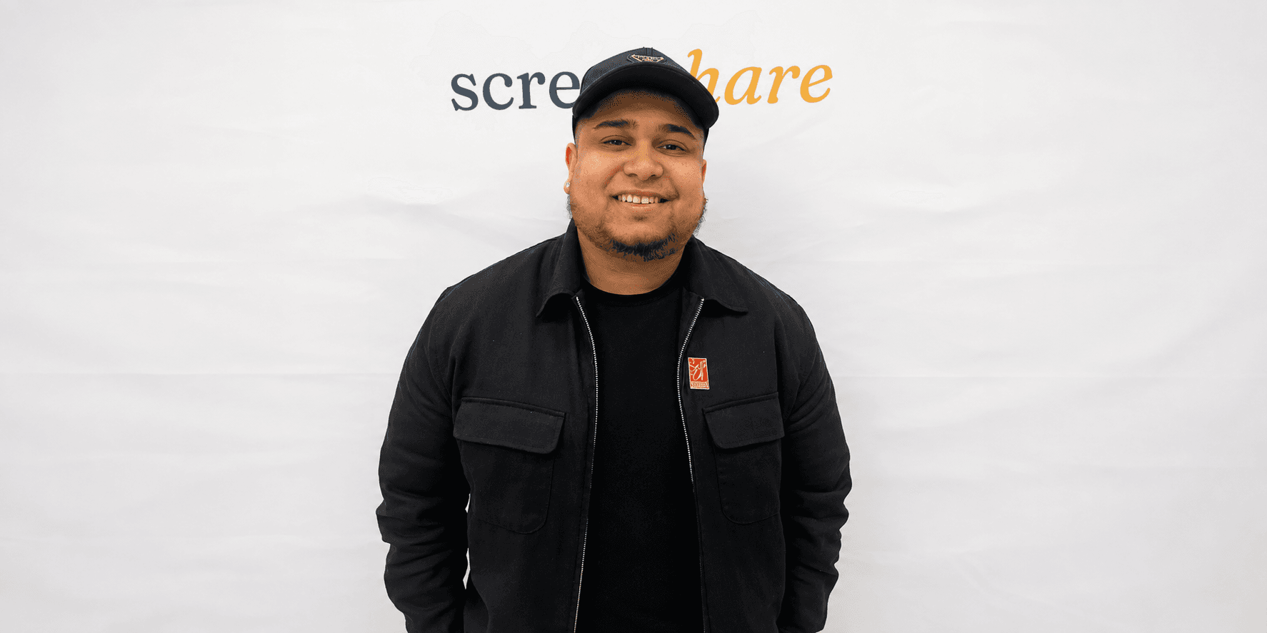Smiling person in black jacket and cap stands in front of a white backdrop