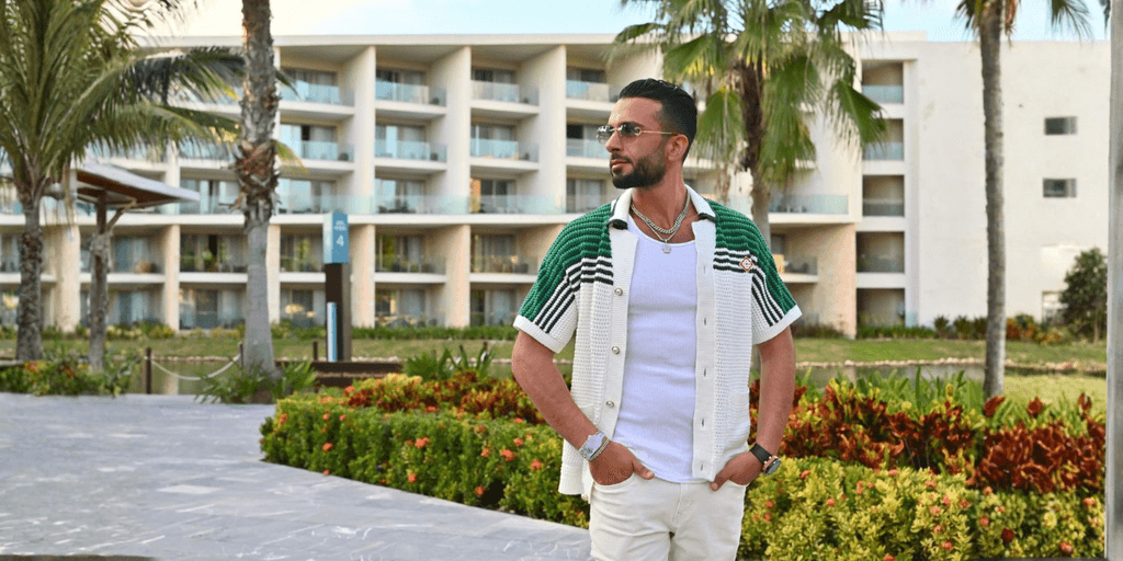 Man in stylish green and white cardigan poses outdoors near a resort.