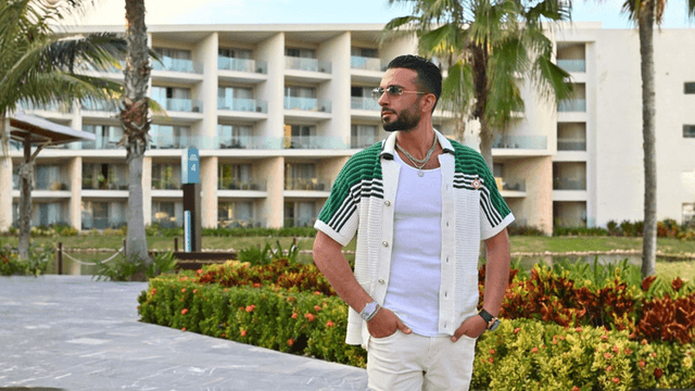 Man in stylish green and white cardigan poses outdoors near a resort.