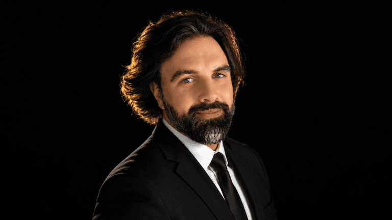 Man in a black suit with a beard, smiling against a dark background