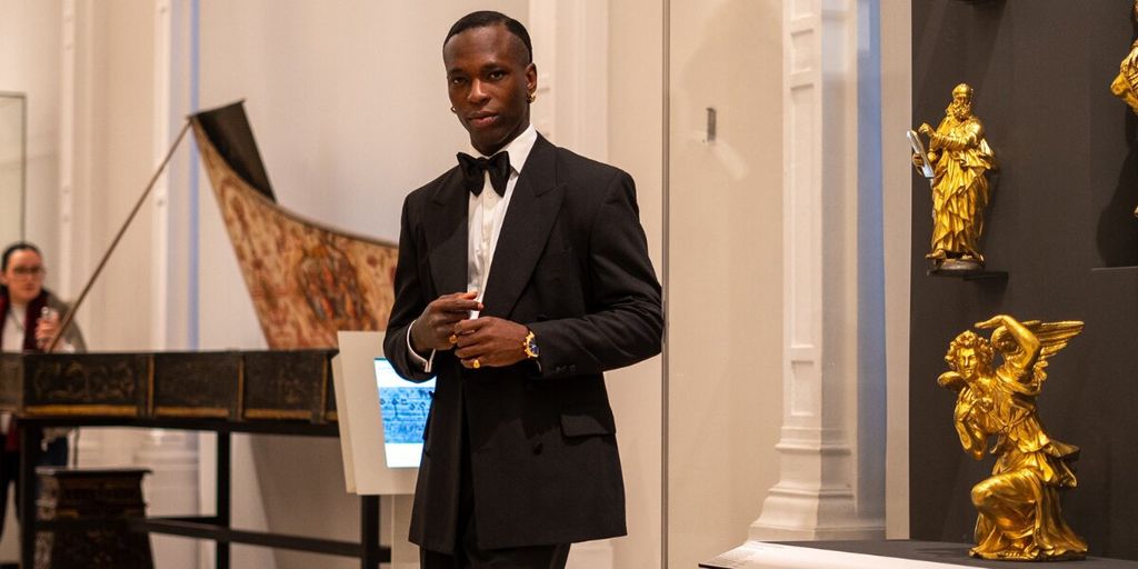 A man in a tuxedo stands confidently in an elegant museum, surrounded by golden sculptures and classical instruments. The atmosphere is sophisticated and formal.