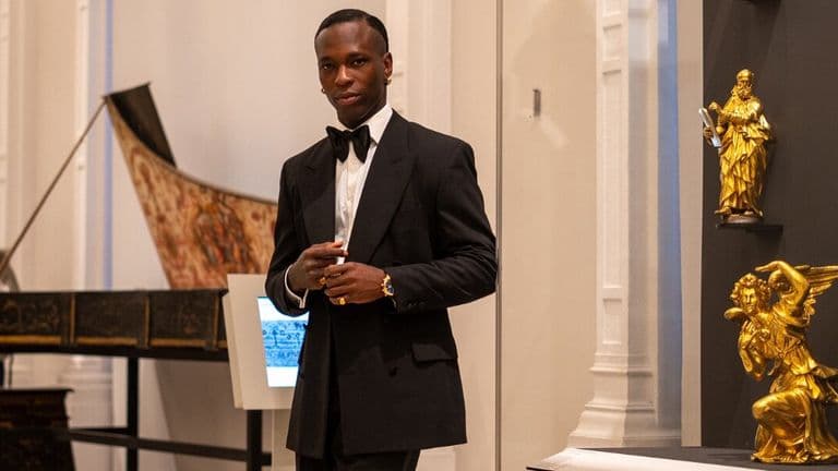 A man in a tuxedo stands confidently in an elegant museum, surrounded by golden sculptures and classical instruments. The atmosphere is sophisticated and formal.