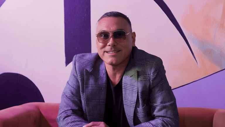 Dylan Garcia smiling in a stylish plaid jacket and sunglasses, seated in front of a colorful, artistic backdrop promoting funk music.