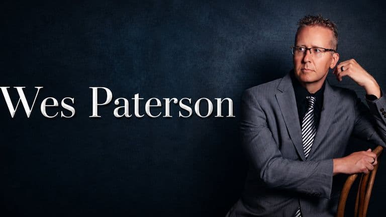 Portrait of Wesley Paterson in a gray suit and glasses against a dark background.