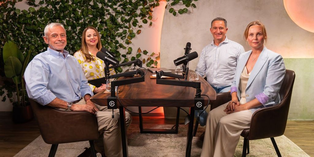 Four people sit around a wooden podcast table with microphones. They smile warmly, surrounded by greenery and soft lighting, creating a relaxed atmosphere.