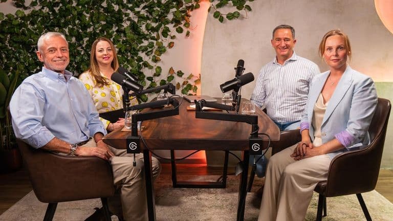 Four people sit around a wooden podcast table with microphones. They smile warmly, surrounded by greenery and soft lighting, creating a relaxed atmosphere.