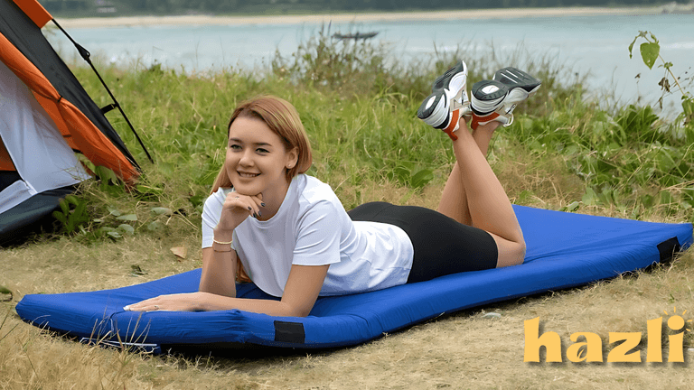 Woman lying on a blue memory foam camping mattress near a tent by a lake, smiling comfortably. Hazli logo visible.