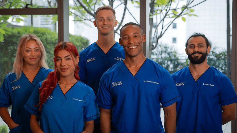 A group of five healthcare professionals wearing blue medical scrubs, standing together in a brightly lit room with plants in the background. They are smiling at the camera.