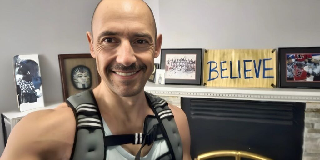 Motivational photo of a person wearing a weighted vest, standing in front of a fireplace with framed artwork and a "BELIEVE" sign on the wall. The person smiles confidently, emphasizing strength and perseverance. The background features a mix of inspiring art pieces and a personal sports memorabilia display.