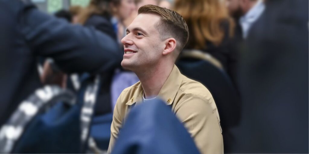 Smiling man in a beige uniform seated in a conference or event setting, surrounded by people, with a focused and engaging expression.